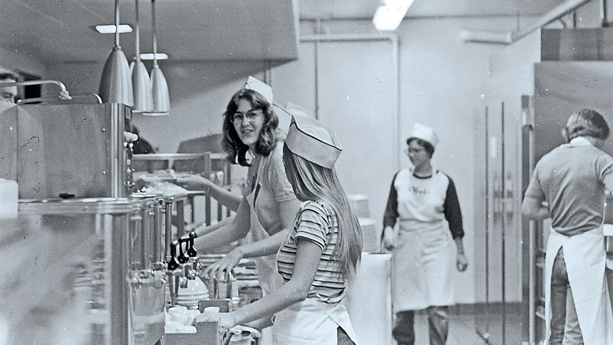 An old black and white photo shows female students serving food at a cafeteria counter.