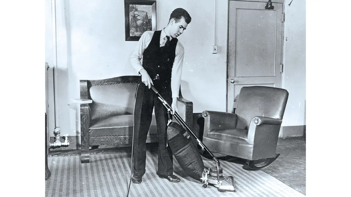 An old black and white photo shows a student diligently vacuuming an area rug while wearing a vest and tie