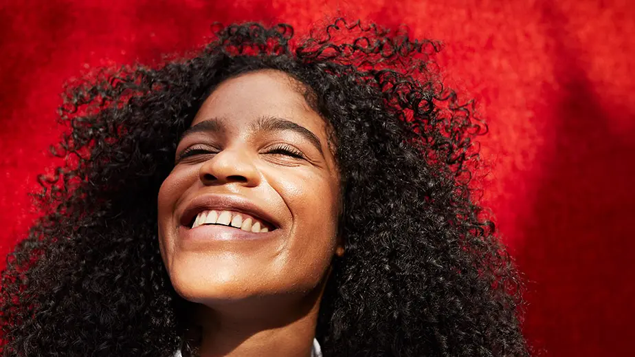 Woman with eyes closed and curly hair smiling with a bright red background