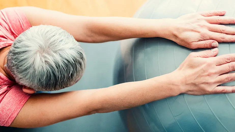 A woman with short gray hair is kneeling on a yoga mat with her hands resting on a teal exercise ball and her arms straight
