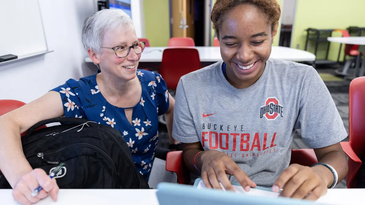 A lady in work clothes and a freshman in an Ohio State T-shirt share a laugh while working on a computer.
