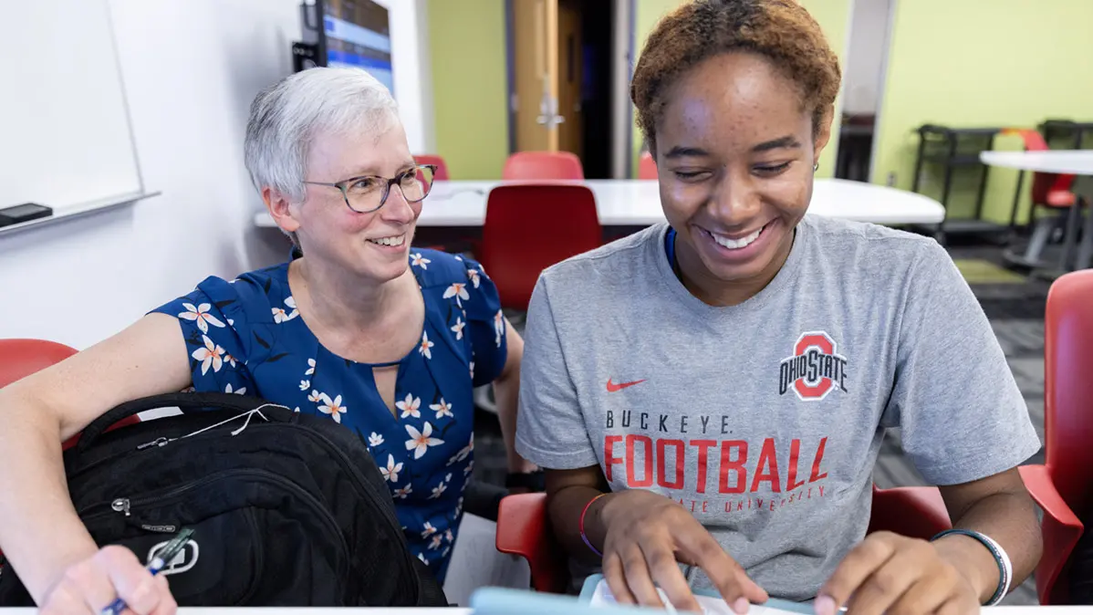A lady in work clothes and a freshman in an Ohio State T-shirt share a laugh while working on a computer.