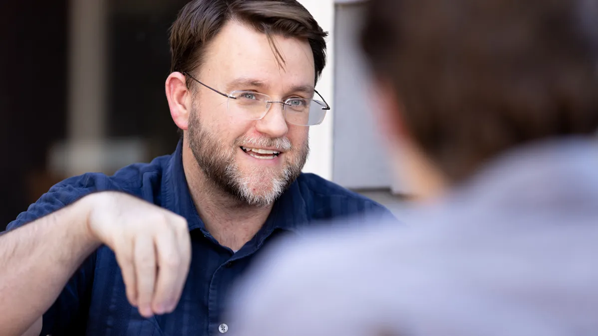 A bearded man wearing glasses meets the eyes of a student who is out of focus as he animatedly explains a thought.