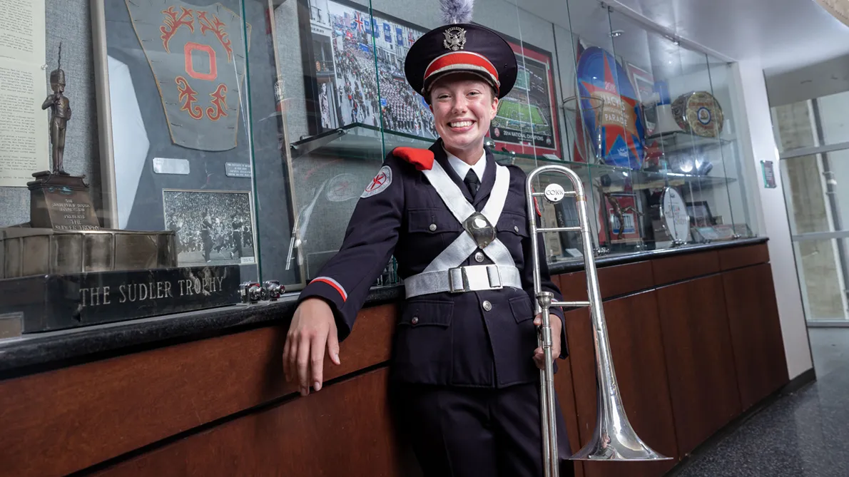 A woman in a marching band uniform smiles brightly as she leans on a counter in front of display cases of TBDBITL memorabilia.
