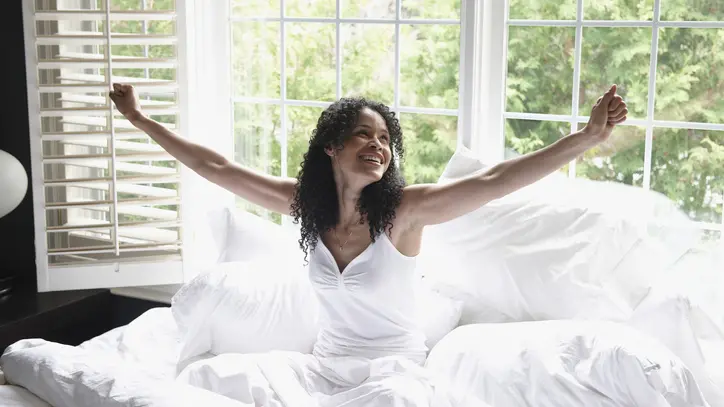 A woman sitting up in bed smiles and stretches as if she’s excited to awaken and take on her day.
