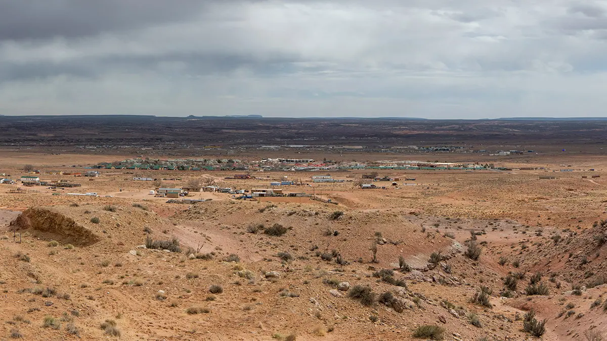 On a cloudy day, a from-far-away look at Chinle shows a small town of mostly single-story buildings with red or green roofs and miles of desert red-brown land stretching to the horizon.
