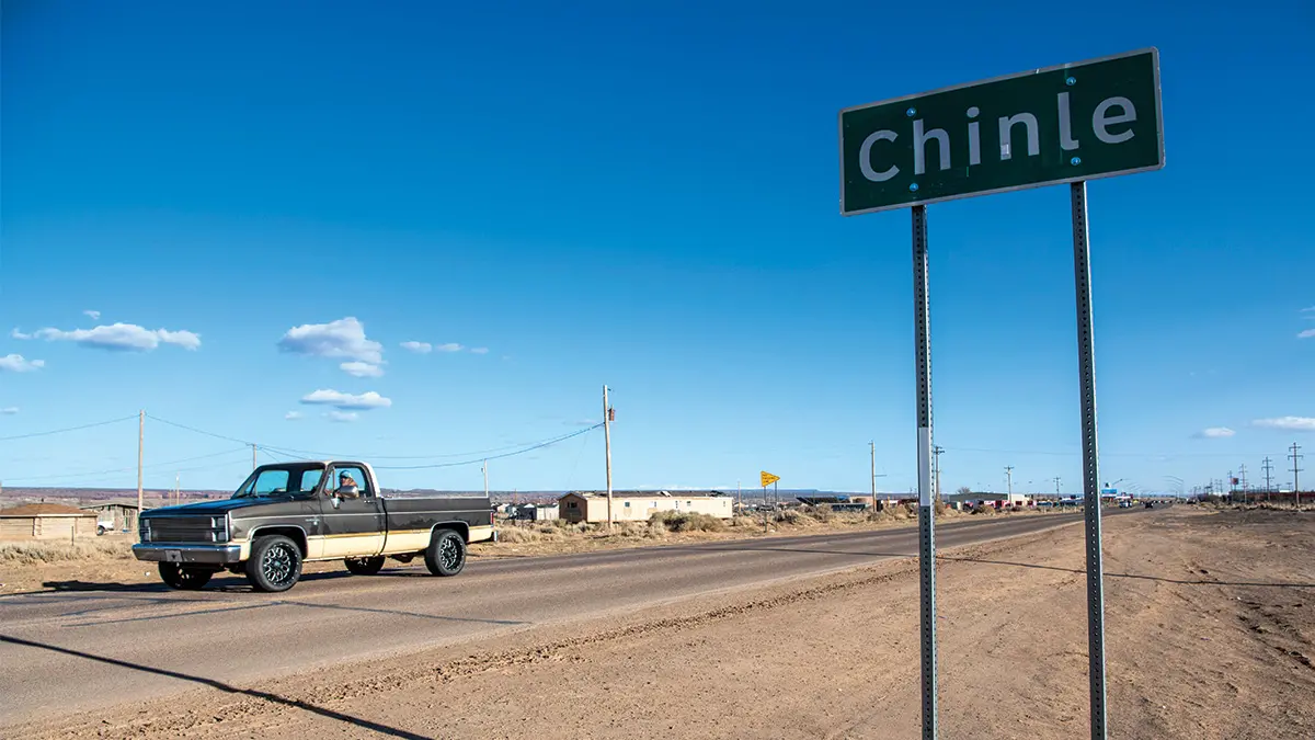 A roadside sign says Chinle. An older model pickup truck drives down the street beside it on a blue-sky day.