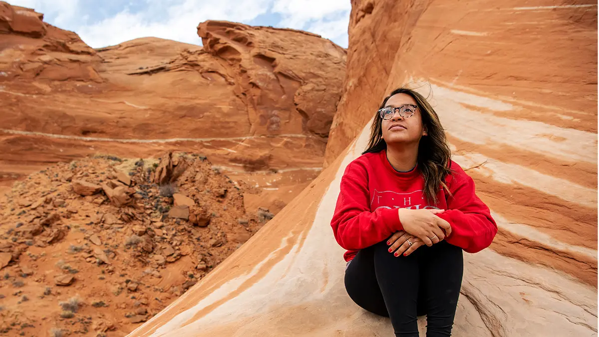A woman in a red Ohio State sweatshirt looks to the sky as she sits on a giant, moon-shaped, orange and cream-colored rock formation. She’s significantly higher than the ground, but behind her, portions of the rock go much higher, off the top of the photo