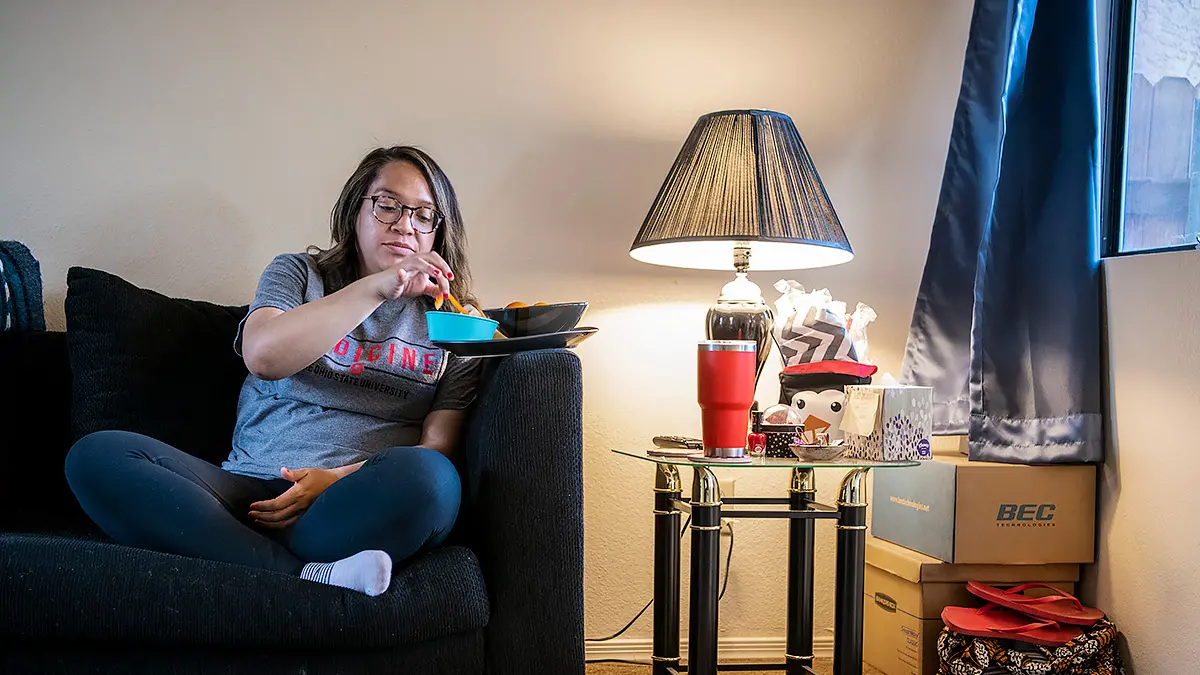 A young woman sits cross-legged on a couch and eats dinner from dishes balanced on the couch's arm.