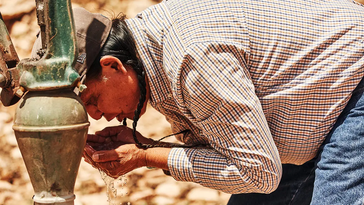 A Navajo Nation man wearing braids and a ballcap bends over to drink water from his own cupped hands, which he just pumped from a green metal well apparatus covered by a thin layer of dirt.