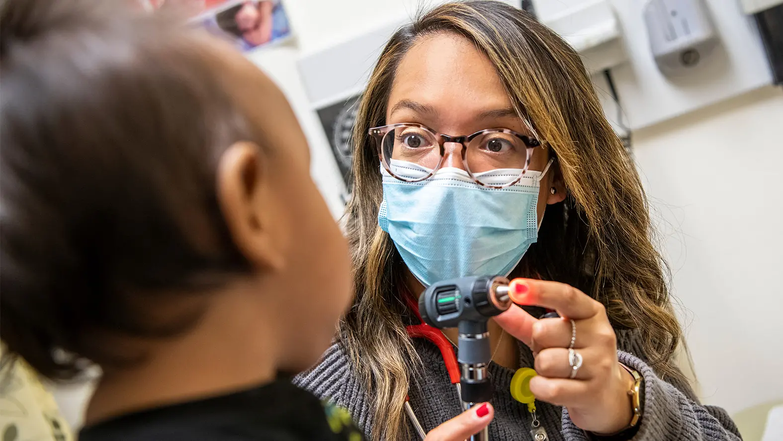 A young doctor of Guatemalan descent wears round tortoiseshell glasses, a surgical mask and a wide-eyed expression as she connects with a toddler. She’s showing the little girl the tool she’ll use to look in her ears.