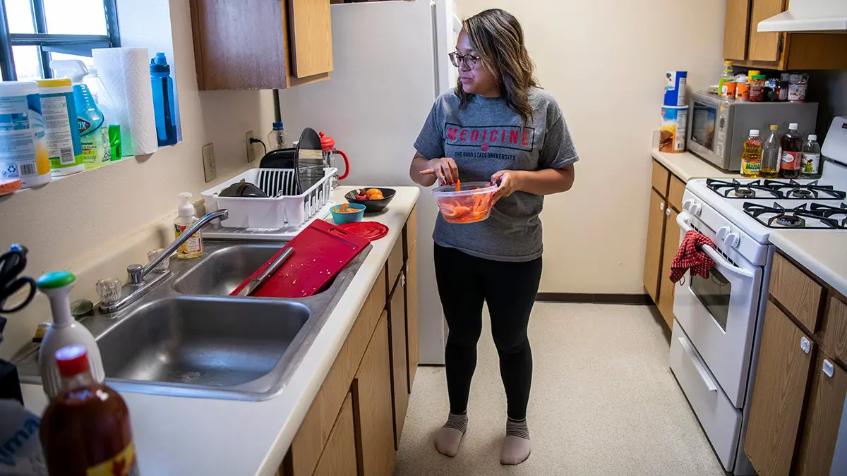 In a narrow kitchen, a young woman in an Ohio State Medicine T-shirt preps bell peppers for future use. On the counter, there is also a bowl of fruit.