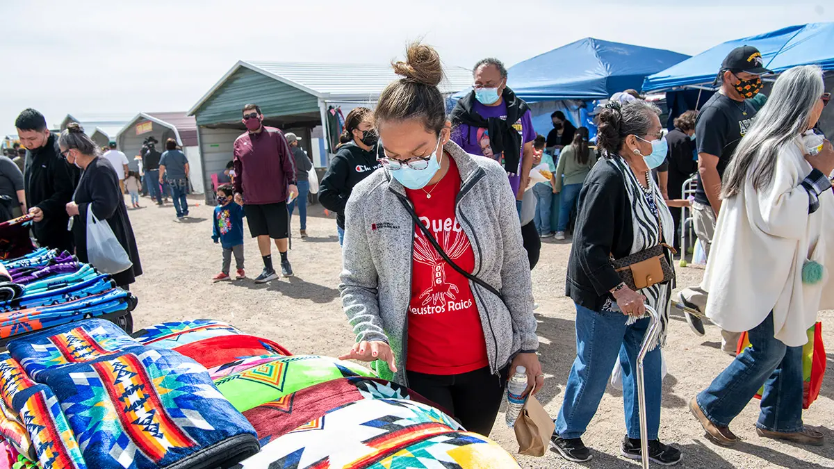 At an outdoors market, a young woman browses colorful, patterned blankets as a stream of Navajo Nation residents pass behind her.