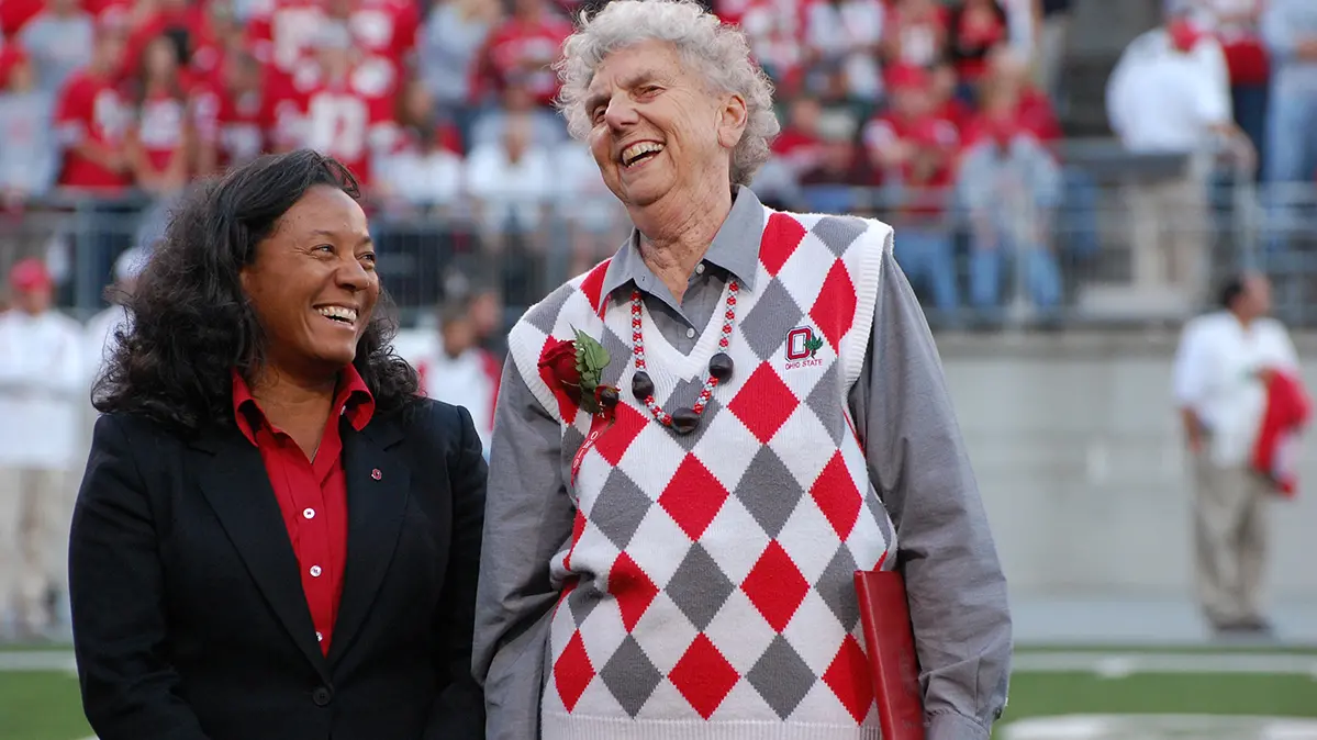 On the field in Ohio Stadium, an older white woman wearing a scarlet and gray argyle vest stands next to a younger black woman. She’s looking at her friend and they are both laughing.