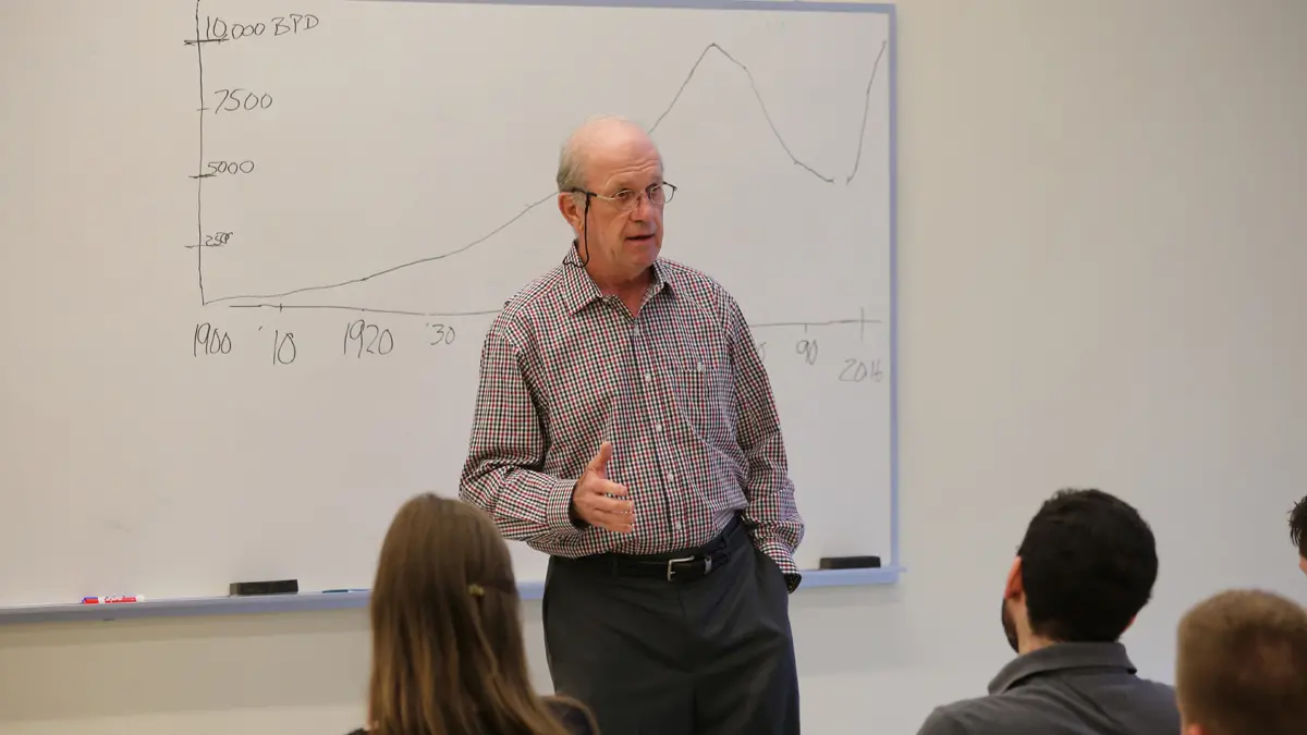 An older man wearing glasses and a checked button-down gestures while teaching a class in front of a white board.