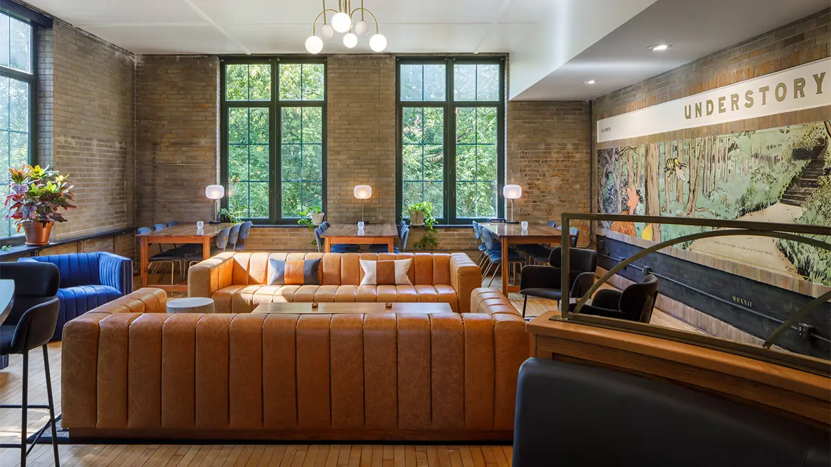 An indoor space has big windows, extra-wide leather-looking sofas and a wall mural showing the Olentangy Trail, steps, botanicals and a large bee. The words above the mural say “understory.”