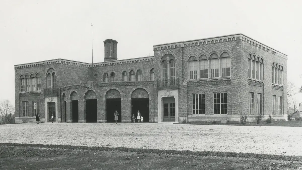 A black-and-white historical photo shows a wide brick school. The center portion rounds outward and there are five archway openings set in that portion. A few adults and a few children stand in front.