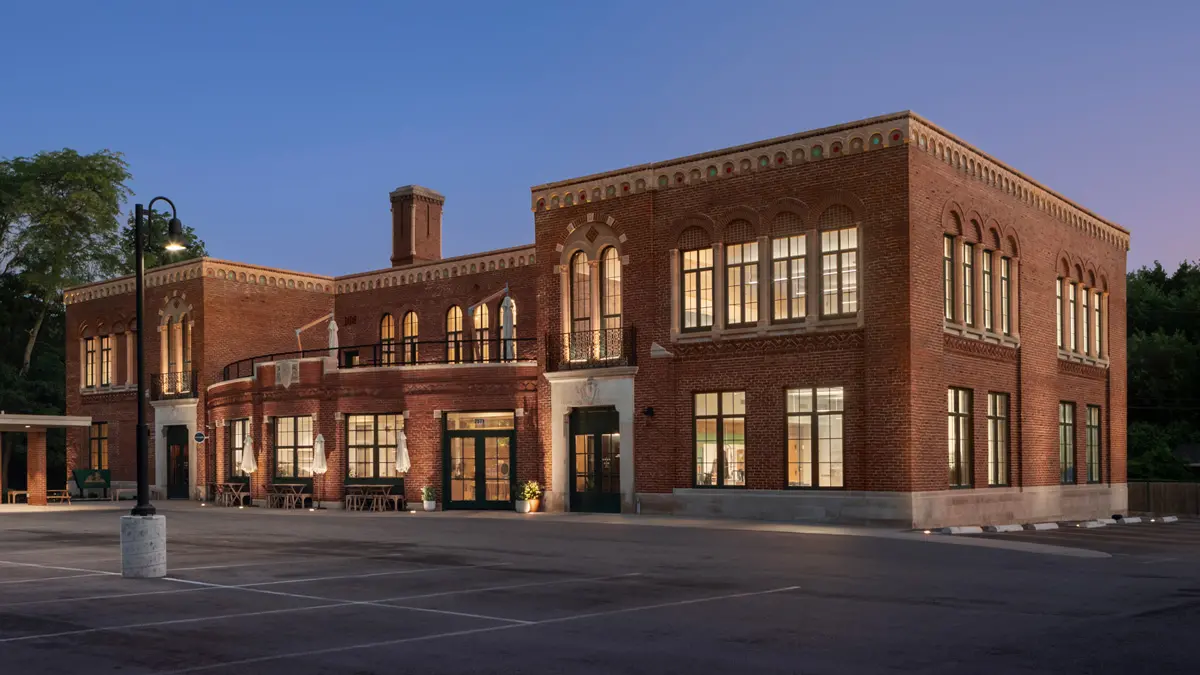 As the sky dims, lights shine from the windows of a wide, squat building whose architectural details include metal railings, arched windows and pale trim accenting the red brick.