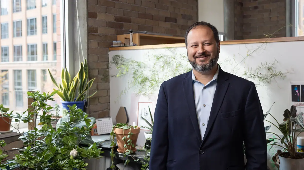 A bearded man in a navy blazer stands smiles broadly in his office, surrounded by plants