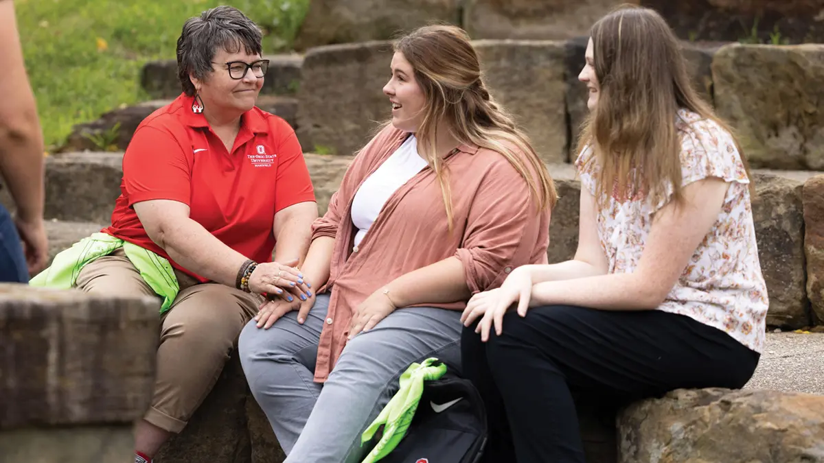 A woman wearing an Ohio State shirt and two students smile at each other in a supportive way as they chat and sit on large stone steps on Ohio State’s Mansfield campus.
