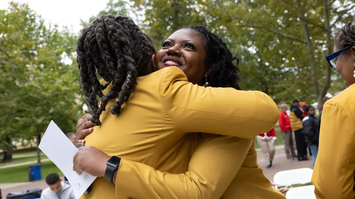 A pair of Black women wearing mustard-colored jackets hug. The woman facing the camera smiles happily as she looks toward the sky.