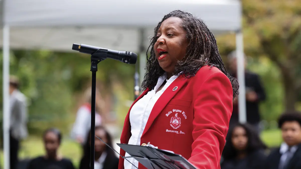 A Black woman intently speaks into a microphone to address a gathered crowd, vaguely seen in the background of the photo.