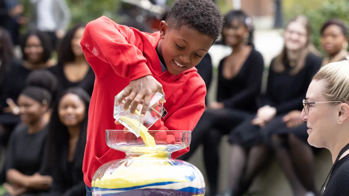 A Black boy wearing a scarlet-colored sweatshirt pours yellow sand from a square glass container into a fancy glass vase wider than he is. The vase is filled nearly to the top by layers of sand of other colors—each layer was poured in by someone else before him.