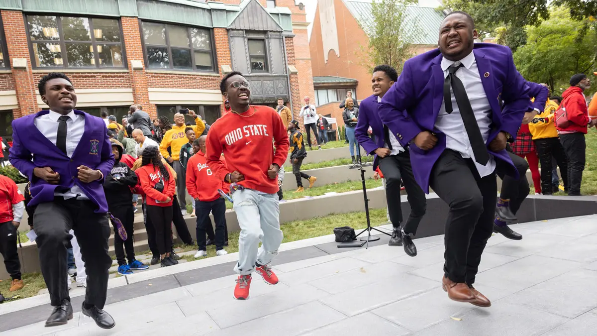 Four young Black men are dancing are in step: Slightly in the air, they all have the same leg lifted and have big smiles or animated expressions.