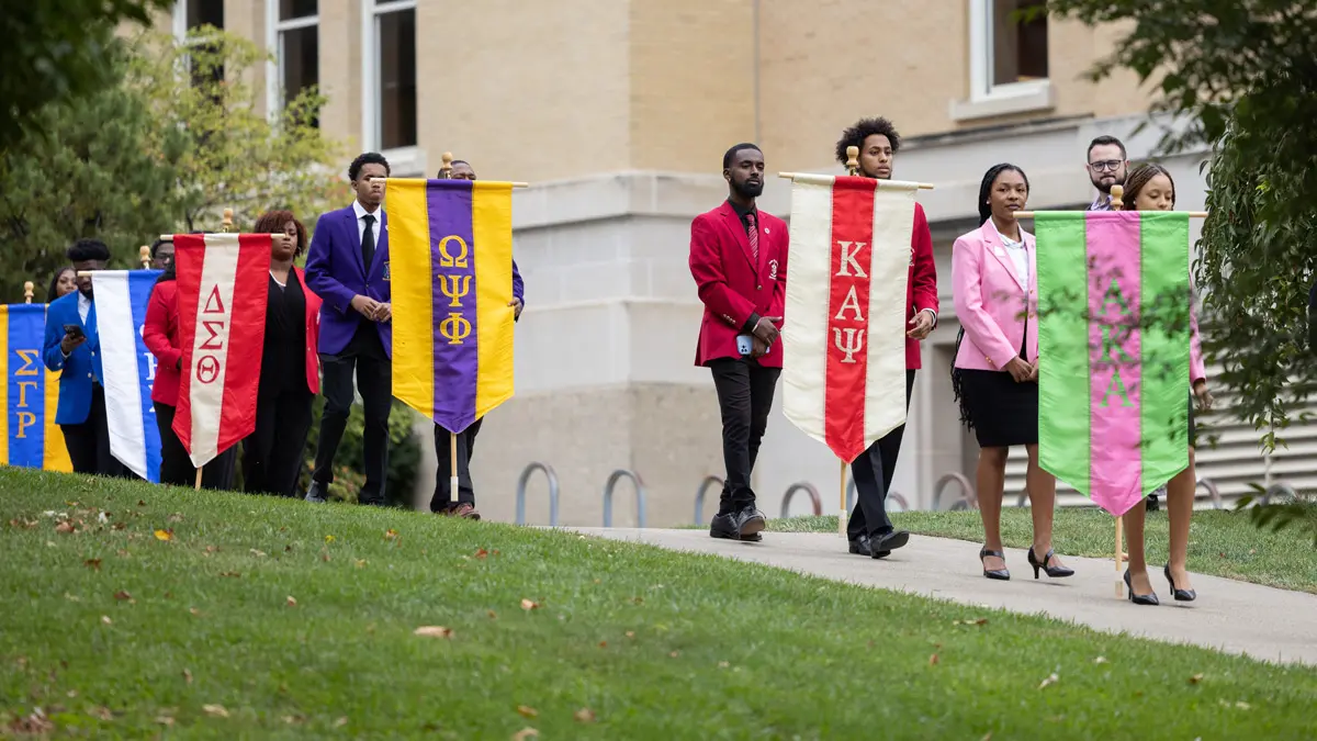 Representatives of the Divine Nine walk on a path toward the plaza. Each pair carries a colorful banner representing their fraternity or sorority.