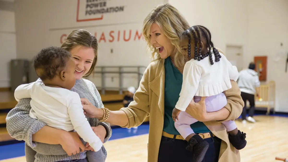 In a gymnasium, two white women each hold a young black child on a hip; the little boy and girl each grasp the shoulder of the woman holding them. Both kids are toddlers. The women are mother and her adult daughter; the mother reaches playfully toward the boy her daughter holds, while the little girl she holds watches.  