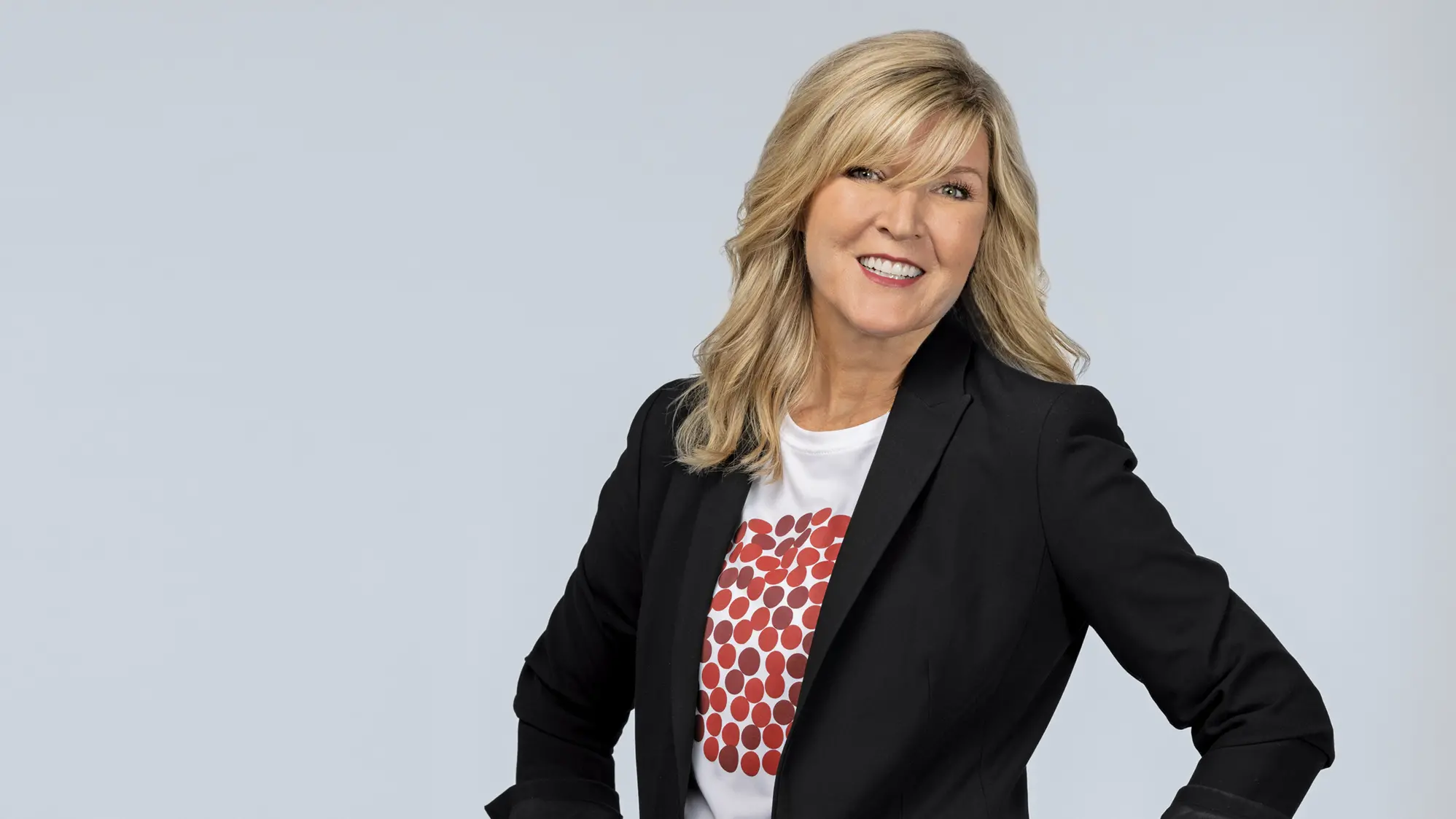 A white woman wears a pepperoni pizza T-shirt, under a black suit jacket, with black pants. She has blond hair, blue eyes, a big smile and stands confidently with her hands on hips.