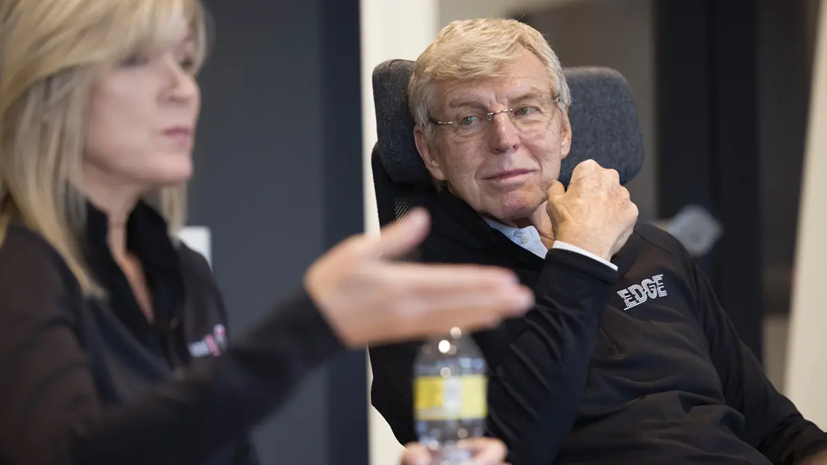 An older white man wearing glasses looks admiringly at his daughter as she makes. Apoint in a meeting. She is slightly out of focus. Both are blond and wear black Donatos shirts. 