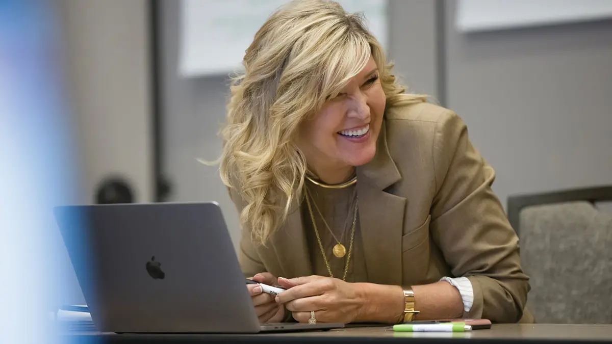 A blond woman in a matching sweater and suit jacket leans forward at a conference-room table as she laughs with someone not in the photo. A Mac laptop sits in front of her. 