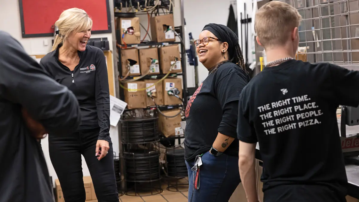 Four people stand in the kitchen at a pizzeria. The two women look like opposites — one is white, thin and tall; one is Black and shorter — but they both have big grins as they chat. The photo doesn’t show the men’s faces. 