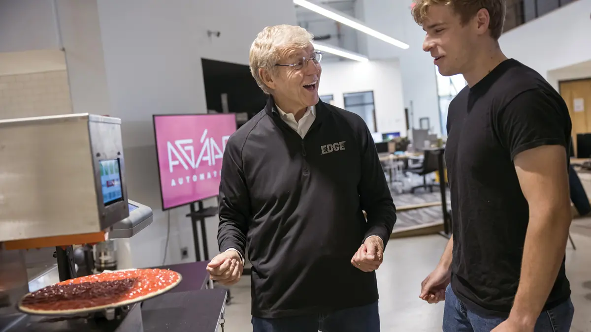 A college student watches an automatic pizza saucing machine as an older man talks to him excitedly.