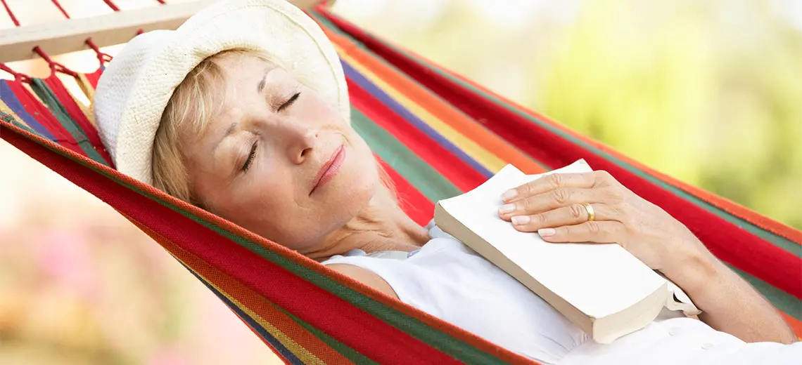 A white woman wearing a sunhat slightly smiles as she sleeps outdoors in a colorful hammock.