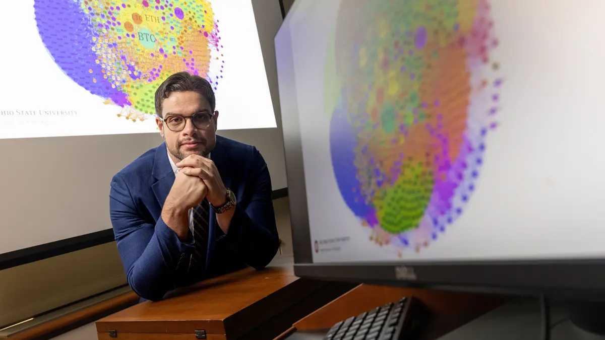 In a classroom setting, a man wearing a suit and glasses looks confident and thoughtful as he leans on a desk between colorful charts.