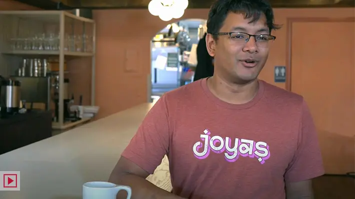 An American man of Indian descent smiles while chatting as he leans on the counter in his restaurant. He looks like he's enjoying himself.