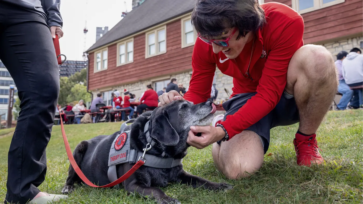 In the grass at an event, a young man wearing red sunglasses and an Ohio State shirt smiles as he kneels to pet a black dog. The dog seemingly gazes adoringly at him as it slightly licks its gray-speckled muzzle and wears a vest that says “therapy dog.”