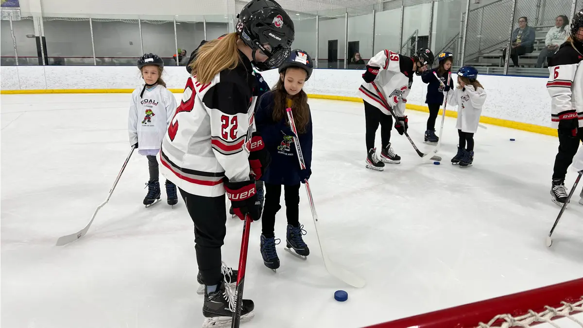 On the ice of a hockey rink, an Ohio State player shows a young girl how to hold her hockey stick and hit a puck. Other players and girls are in the background
