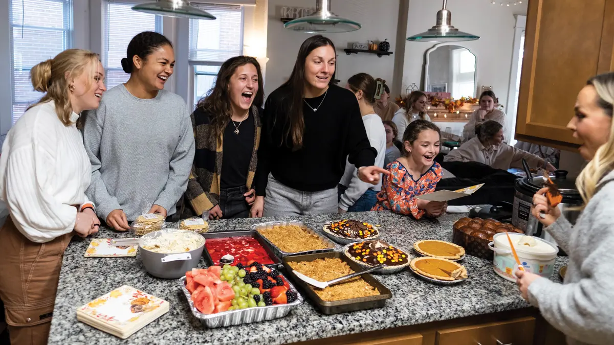On one side of a kitchen island loaded with fruits, pies and rectangular pans of more desserts, four young women and a girl laugh while looking at the young woman on the other side of the island. She holds a knife in one hand and in the other, the cover for that knife, which is caked in chocolate.