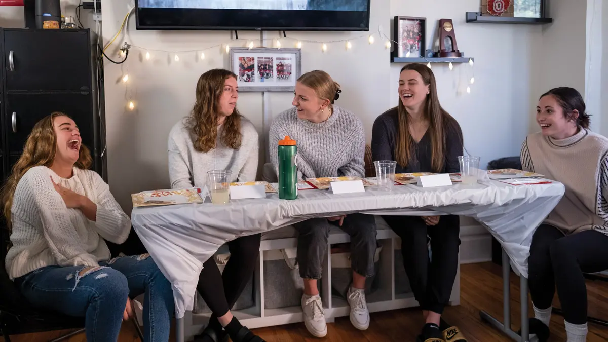 Five young women wearing sweaters sit at a table with soon-to-be-filled paper plates. The woman on the left is laughing so hard she has her hand on her chest. Ohio State signs hang on the wall behind them and a TV mounted above their heads shows a crown of people waving, as if they’re looking at you.
