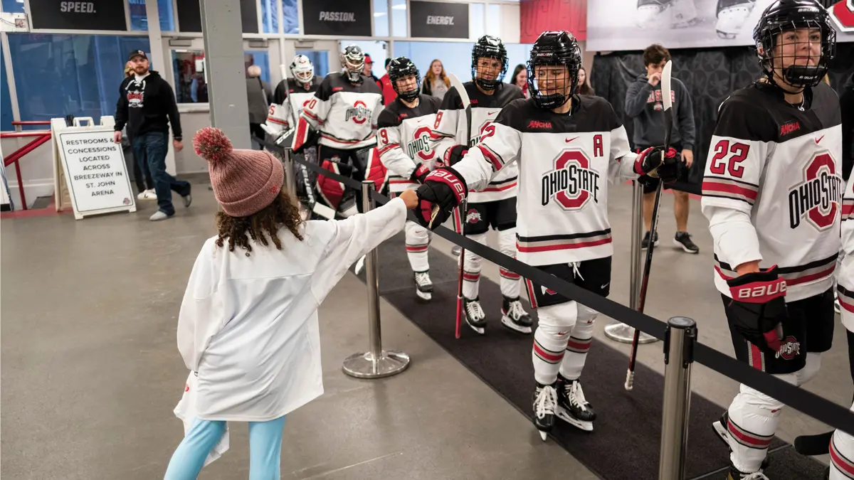As a line of women’s ice hockey players walk in uniform and skates on a roped-off mat toward the ice rink, a young fan reaches to greet a player. The girl, seen f  stocking cap topped with a yarn ball. The player smiles as she extends her gloved hand.