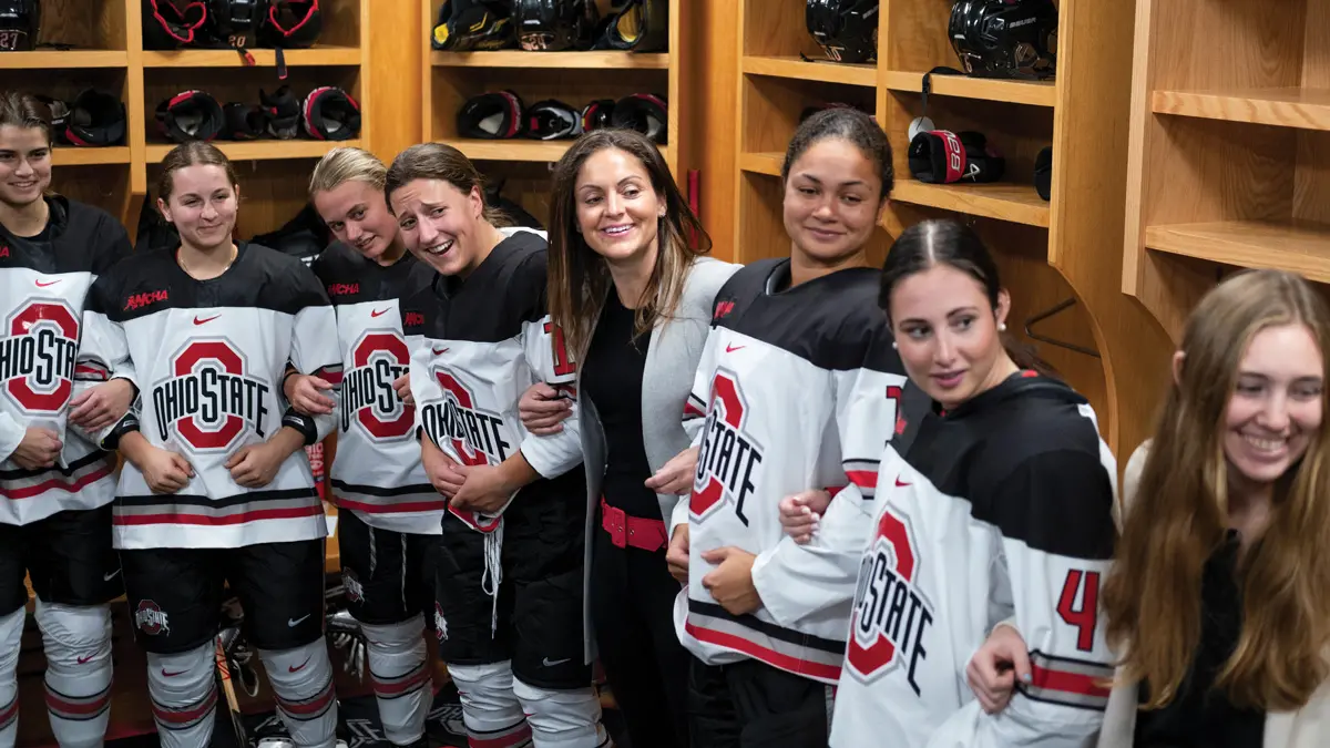 Eight women stand with arms linked along the edge of a locker room. They’re all looking at someone off camera and have varying levels of smiles. Most are players; the head coach is among them. Behind them are open-faced lockers: nice wooden shelves that hold hockey helmets and other gear for each player.