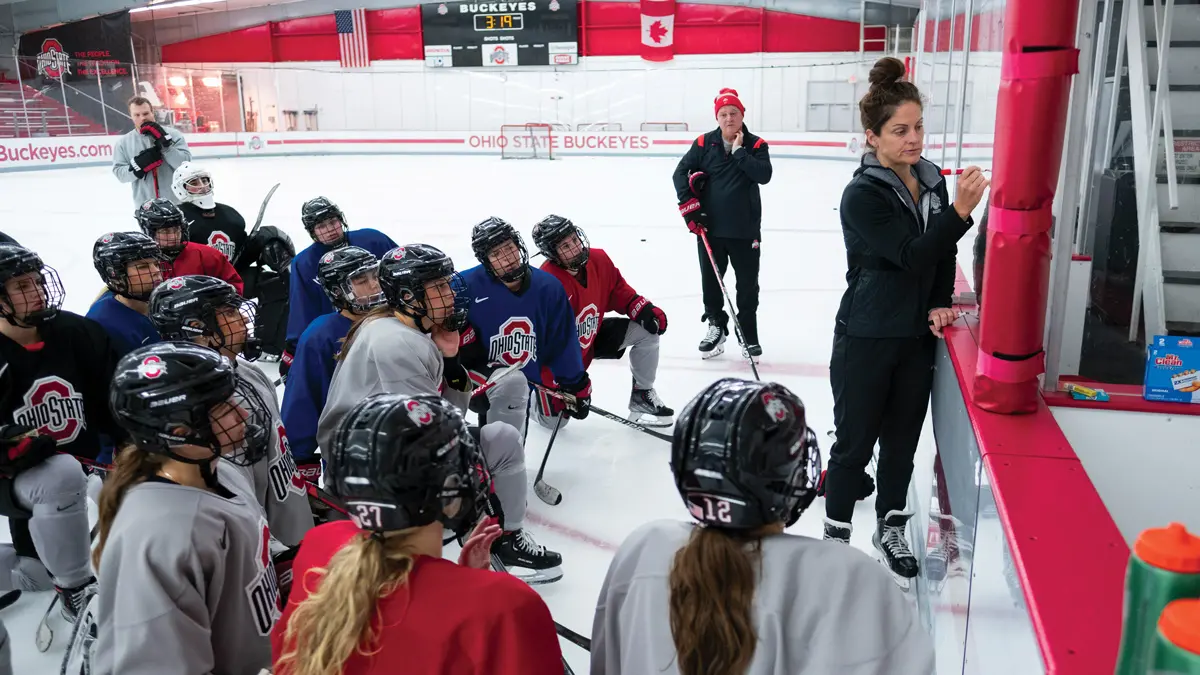 At least 15 ice hockey players crouched in an ice rink intently watch a woman using a dry erase marker to draw on the plexiglass surrounding the rink. She is their head coach, and two male coach assistants stand on the periphery.