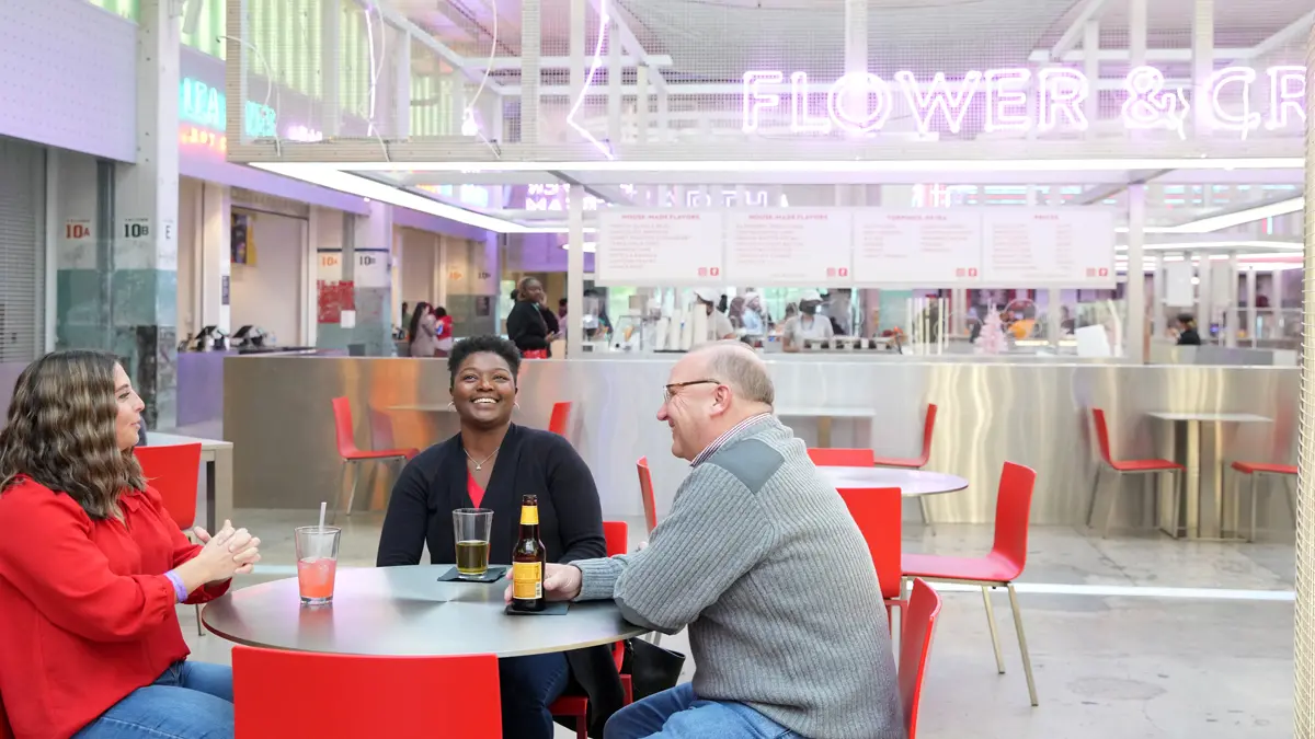 In a restaurant with stainless steel counters, tables with red chairs and a gray concrete floor, three Buckeyes sit together. The woman on the left talks with her hands folded; the woman next to her laughs while looking up, and the man laughs while holding a bottle of beer.