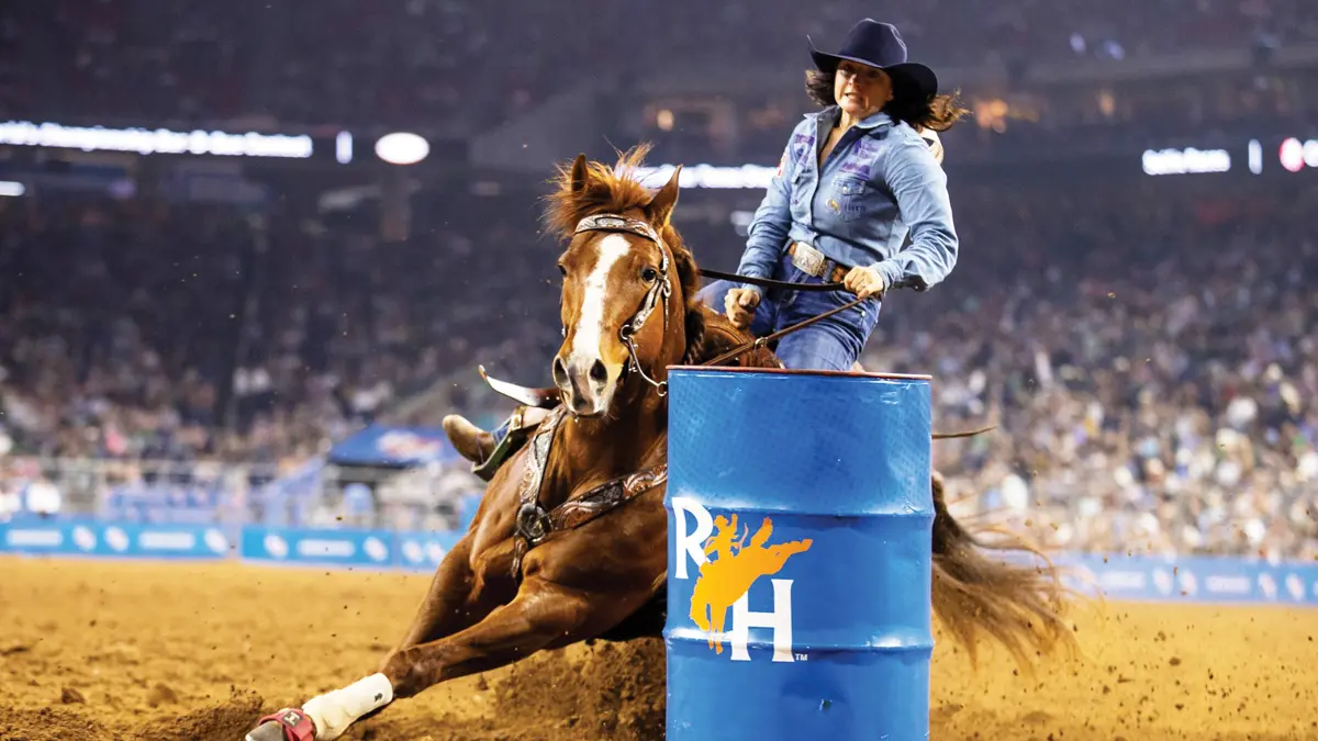 A woman in a cowboy hat rides a horse looping so fast around a barrel, it’s leaning at a 45-degree angle. They’re on dirt in a stadium and the crowd in the stands in the background is vague but huge.