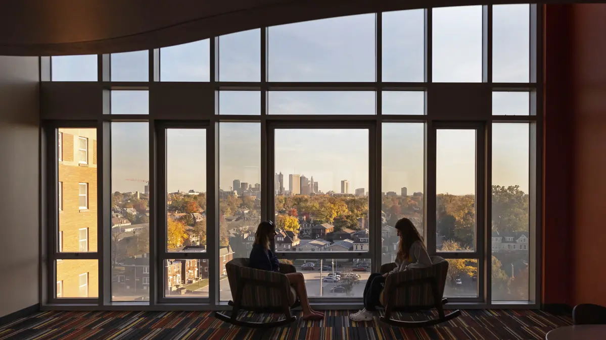 Two young women sit in padded rocking chairs in front of a wall of windows that make them look like no more than dark silhouettes. The frames in the wall of windows have an architectural look and in the distance, you can see the skyscrapers of downtown Columbus.