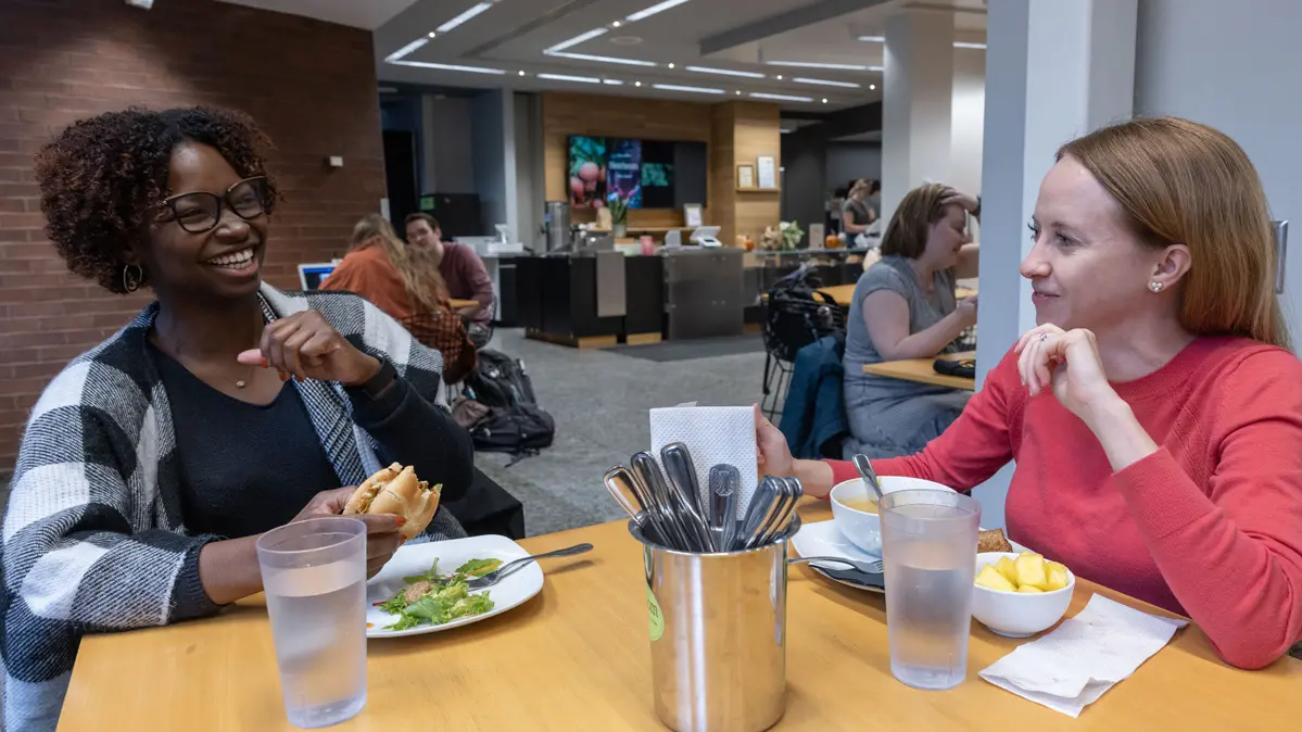 In a restaurant space with an order counter and diners in the background, two young women eat lunch at a wooden table with food in front of them. The black woman laughs, wears glasses and holds a sandwich; the white woman smiles at her and momentarily ignores her soup.