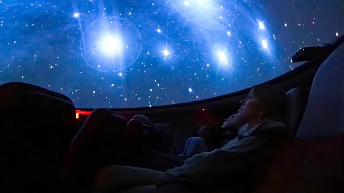 Students sit in seats looking up at a night sky projected onto the domed ceiling of a planetarium. The sky is blue with large and tiny white stars and casts blue light on the students’ faces.